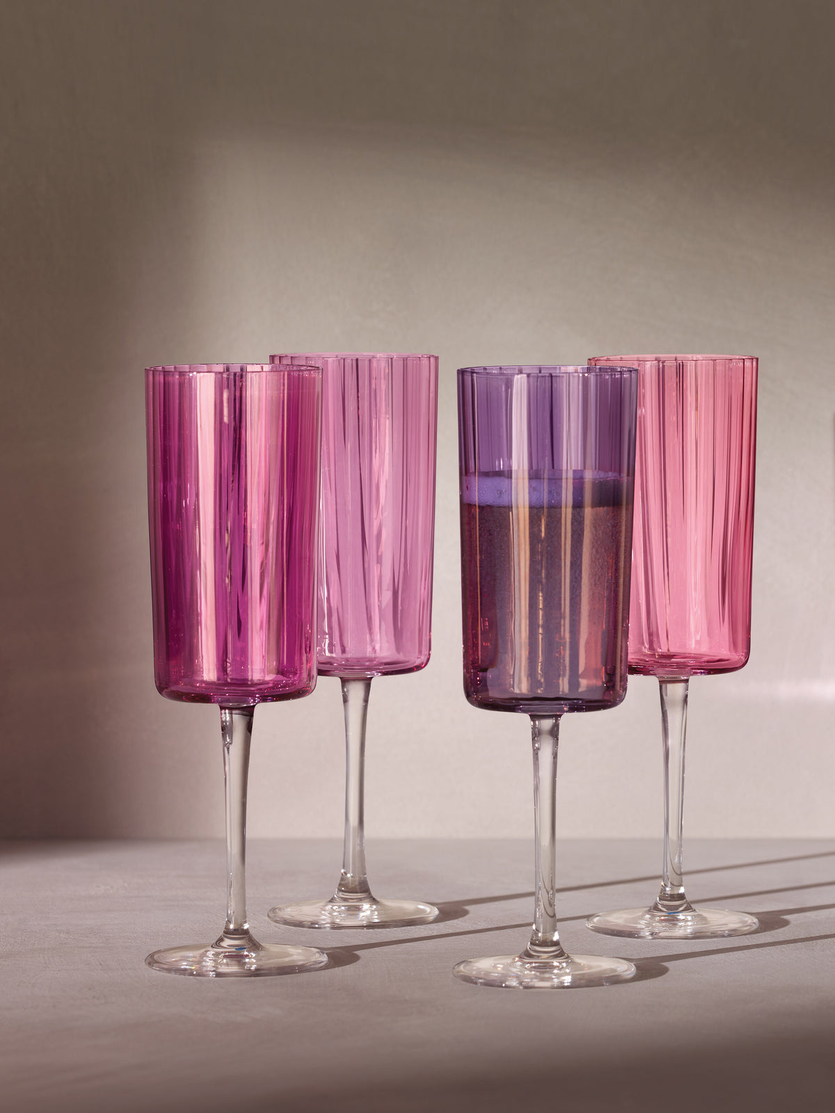 Four tall Champagne Flutes by lsa-international, featuring a pink-tinted hue and vertical textured grooves, stand on a neutral surface. One glass is filled with sparkling liquid while the others remain empty. Soft, diffused light casts gentle shadows across the scene.