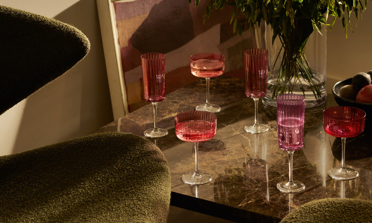 Six vintage-style pink and red lsa-international Champagne Flute glasses are displayed on a dark marble table, accompanied by a vase of green foliage and a fruit bowl in the background, positioned beside green textured chairs and a framed abstract art piece.