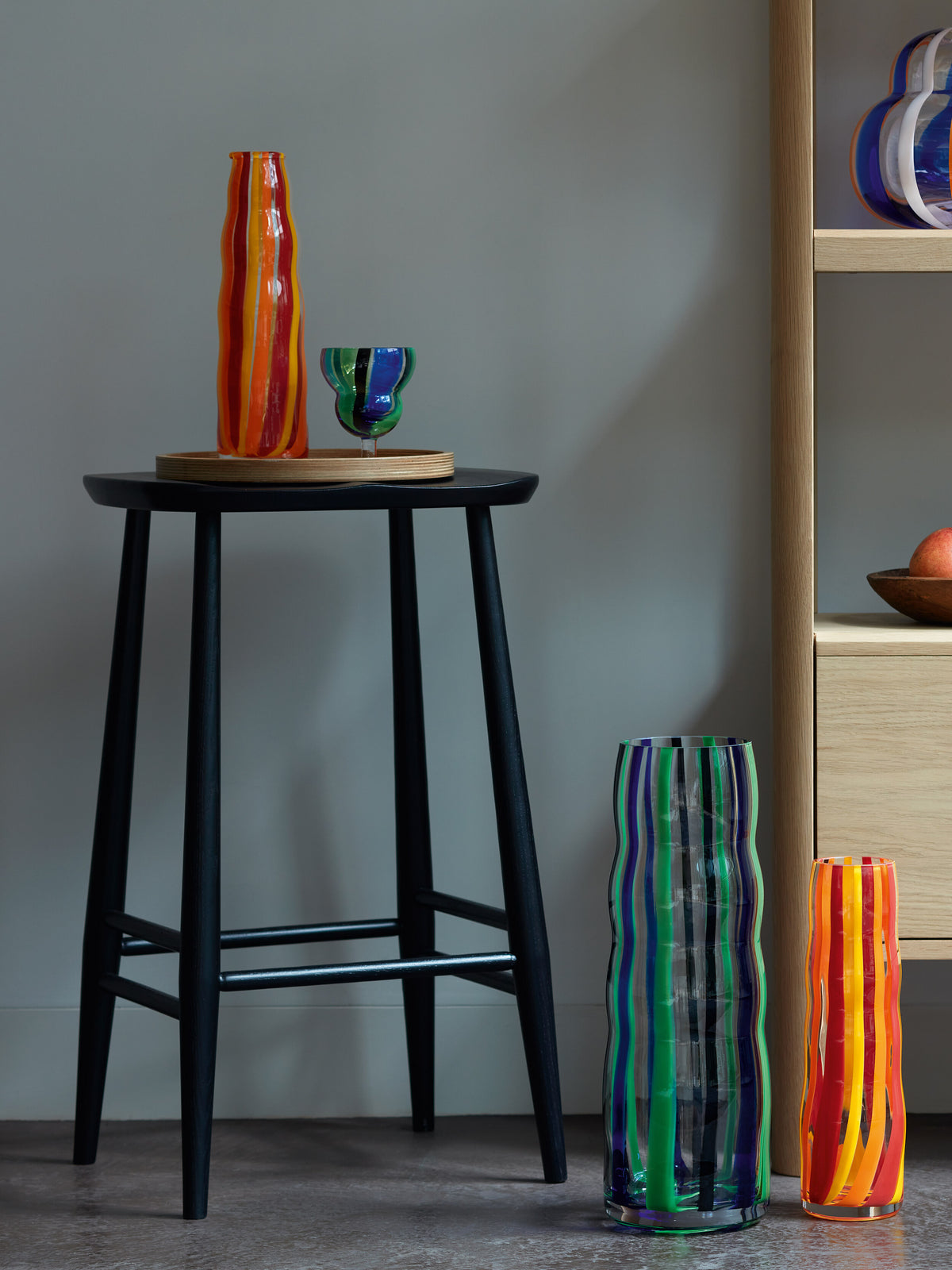 A collection of lsa-international Carafes in colorful striped glass, available in various sizes, sits on a black stool and a wooden shelf in a modern minimalist room, their folk-inspired patterns bringing artisanal charm to the elegant setting.