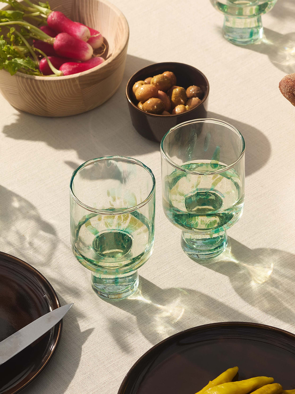 Two partially filled lsa-international Wine Glasses, crafted from green-tinted recycled glass with a subtle speckled pattern, rest on a beige tablecloth surrounded by a bowl of radishes, a small bowl of olives, brown plates, and yellow peppers. Sunlight casts gentle shadows across the table.