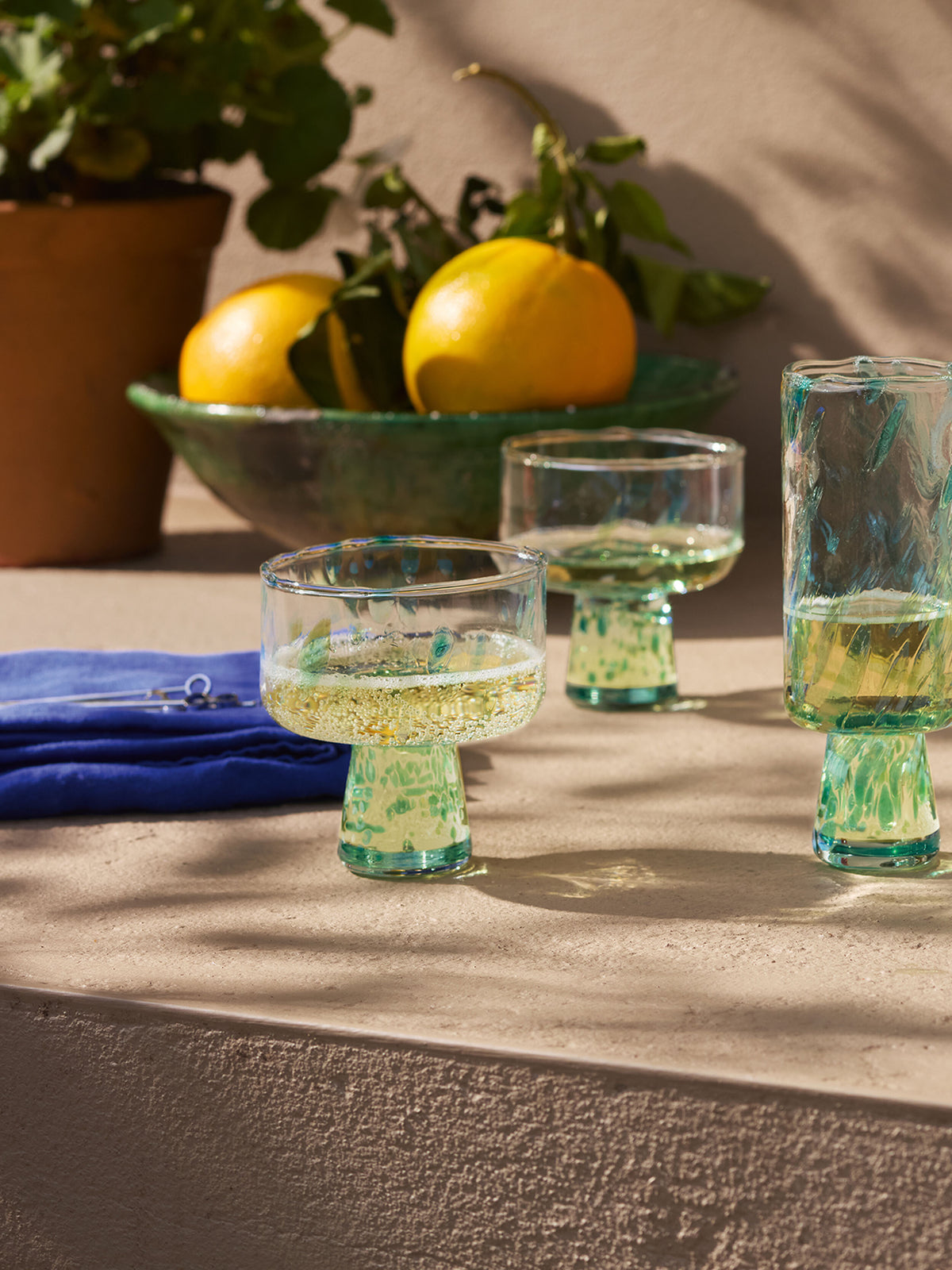Three textured lsa-international Cocktail Saucers, each filled with a yellow beverage, rest on a sunlit stone surface near a blue napkin. In the background, a green bowl holds two yellow fruits with leaves beside a potted plant.