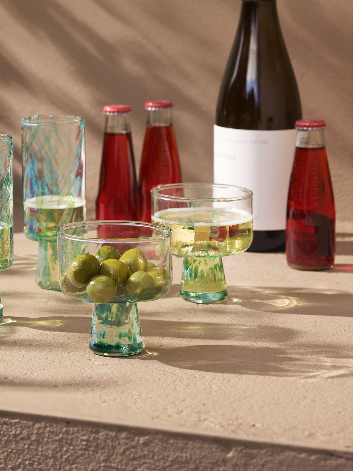 Two goblets with green olives, glasses of white wine resting on lsa-international Cocktail Saucers, a wine bottle, and three red soda bottles are arranged on a beige textured surface in sunlight.