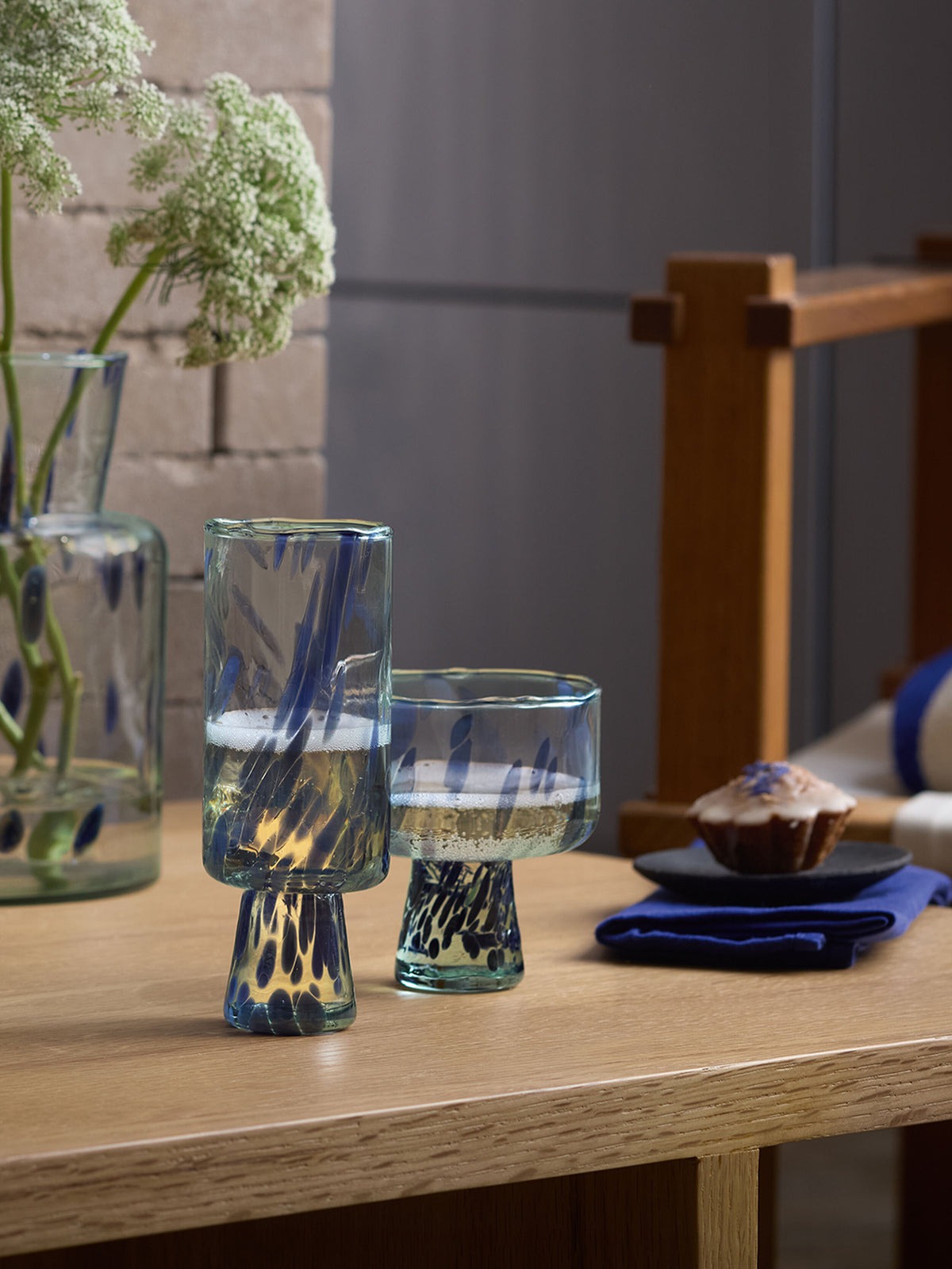 Two distinctive, blue-streaked Cocktail Saucer glasses by lsa-international sit on a wooden table next to a small muffin on a blue napkin. In the background, white flowers in a glass vase and part of a wooden chair are visible.