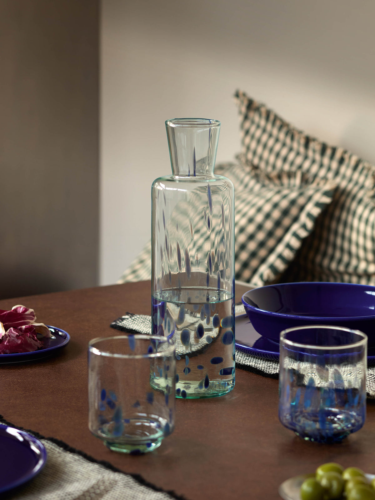 A clear recycled glass Carafe by lsa-international, featuring a blue speckled pattern and partially filled with water, stands on a set table with matching glasses, blue dishes, and checkered pillows in the background.