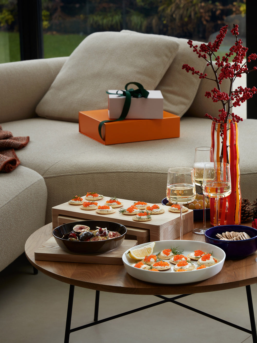 A cozy living room scene with a beige sofa, festive treats on a table set with contemporary tableware, drinks, lsa-international Starter/Pasta Bowls filled with snacks, a vase with red berries, and wrapped gifts in the background.