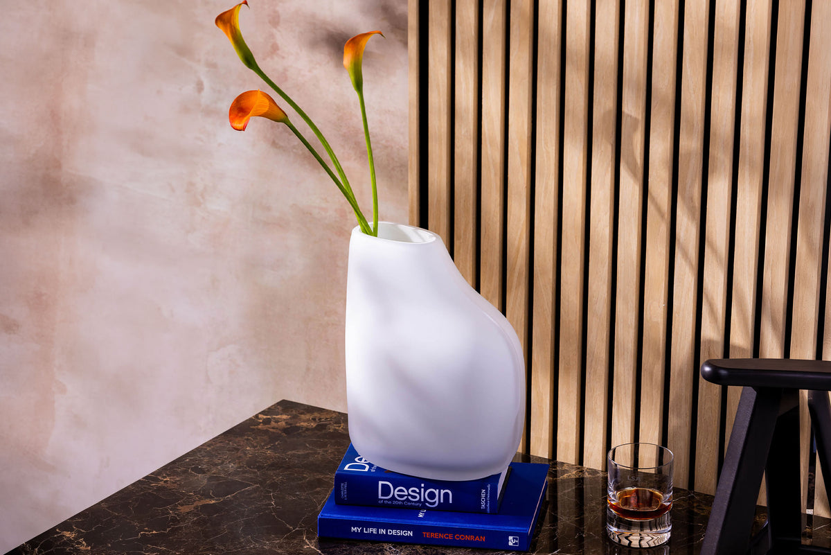 A modern white vase with orange calla lilies sits on two blue books atop a marble surface, near a glass of dark liquid and a wooden slat wall.