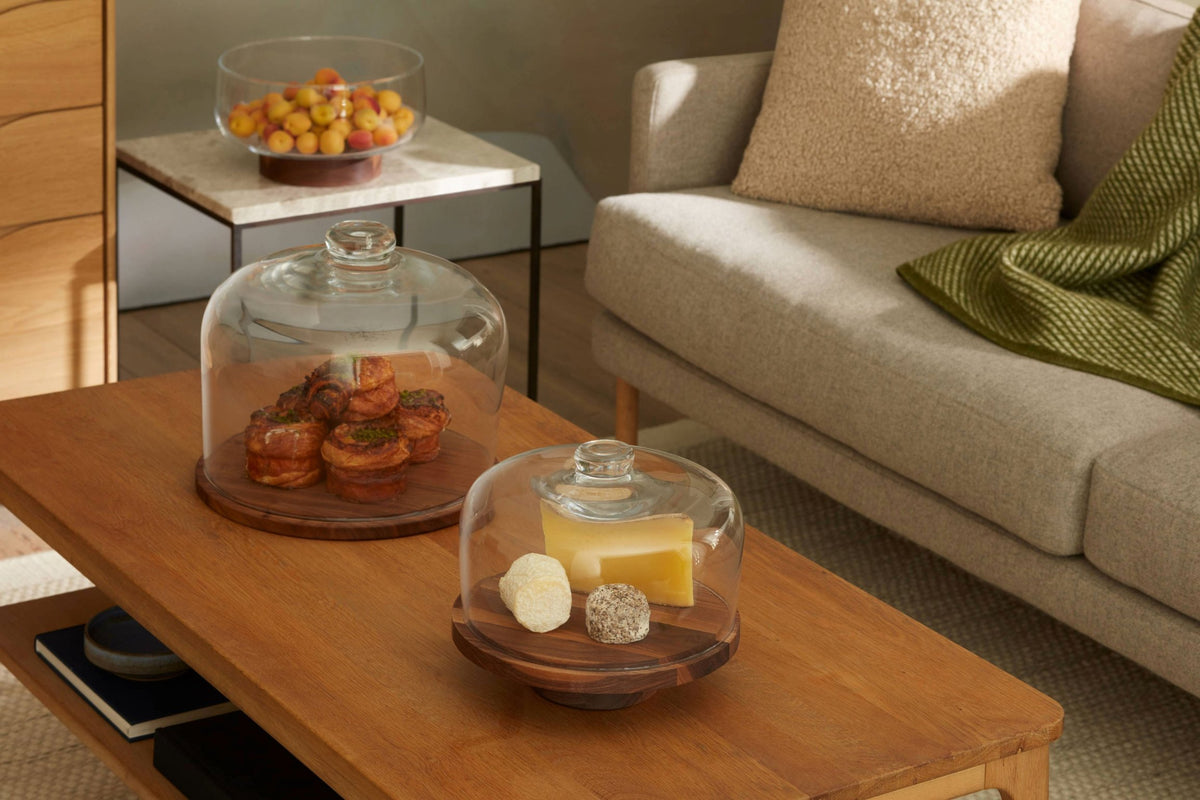 A wooden coffee table with pastries, cheese, and fruit under glass domes. A gray sofa with a green blanket and beige pillow is in the background. A bowl of fruit sits on a side table.