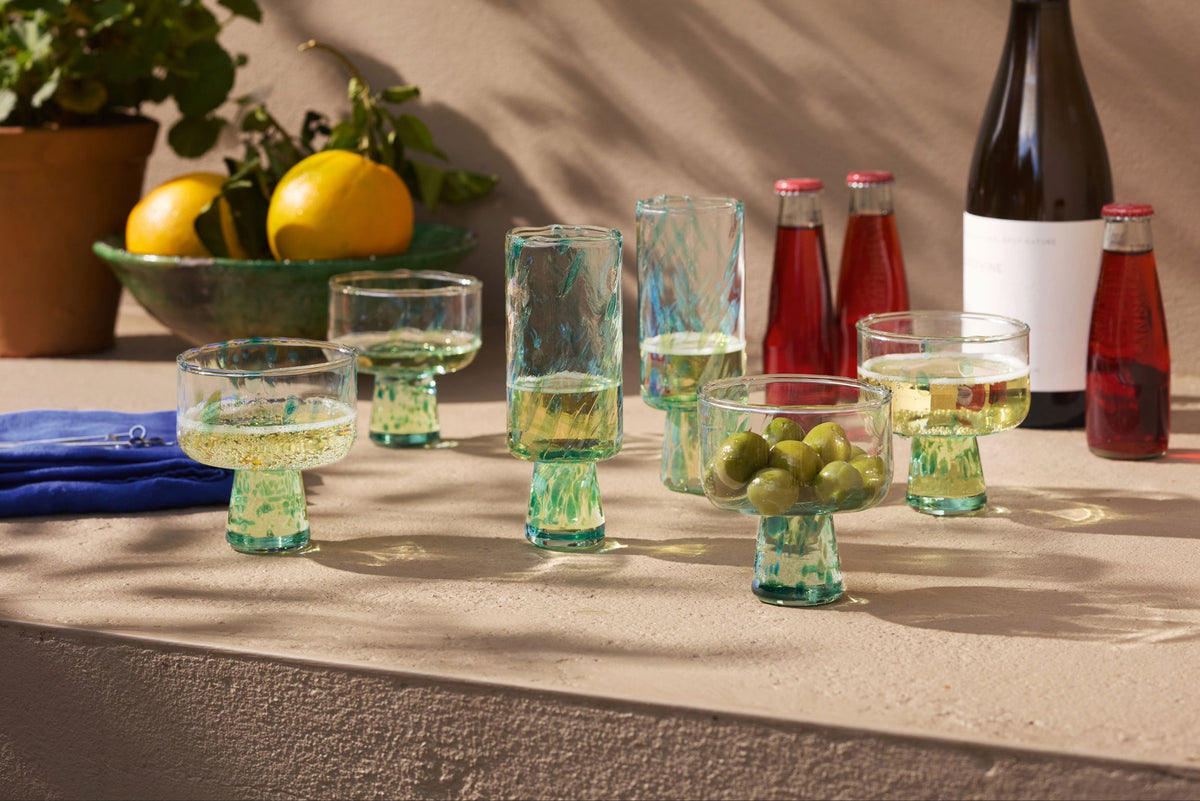 Green drinks and green olives in textured glass cups sit on a stone tabletop. There are patterned glasses, bottles of red drink, a wine bottle, a blue napkin, and a bowl of citrus fruits with leaves in the background.