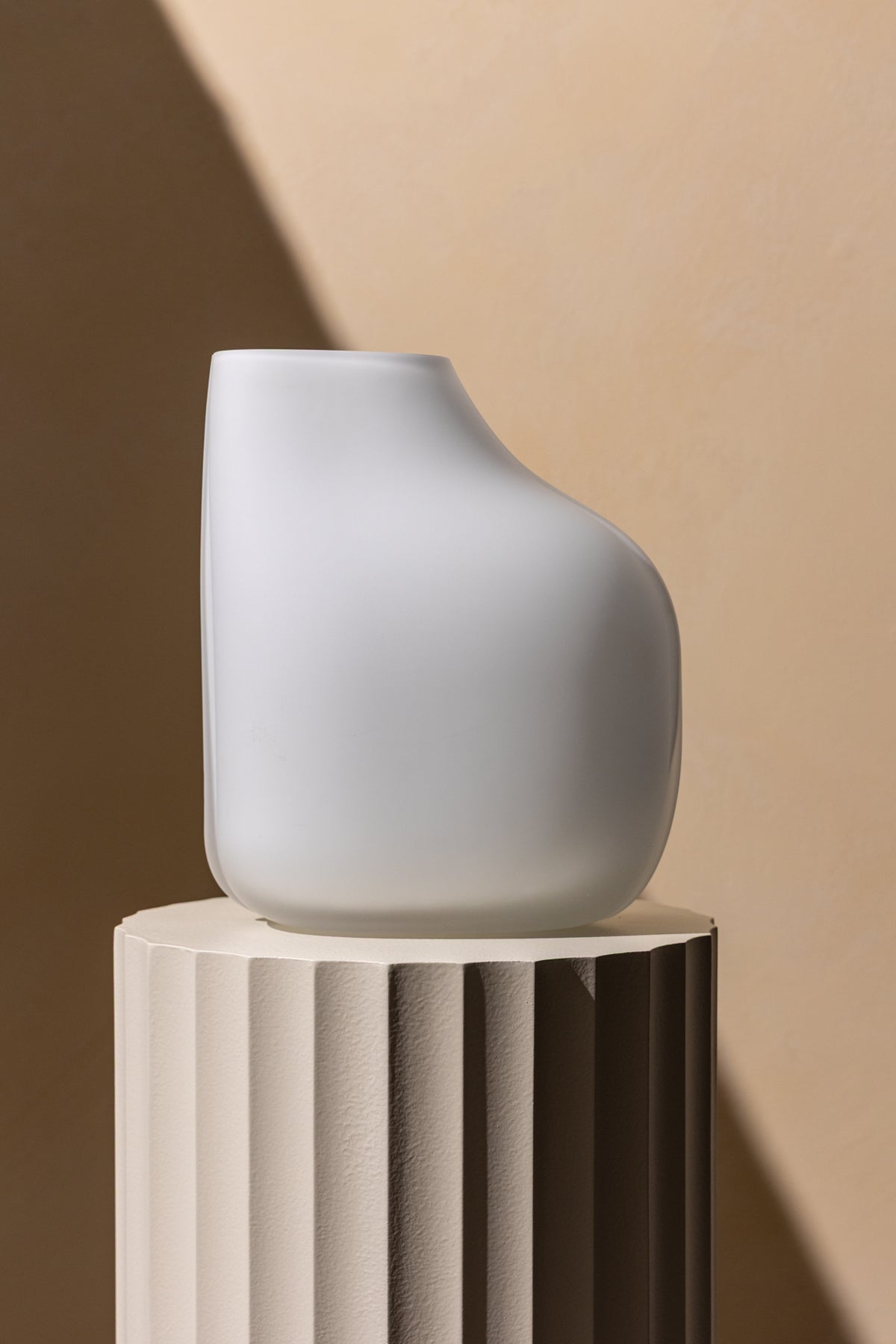 A modern, asymmetrical white vase with a matte finish sits on a textured, fluted pedestal against a beige background with dramatic lighting and shadows.